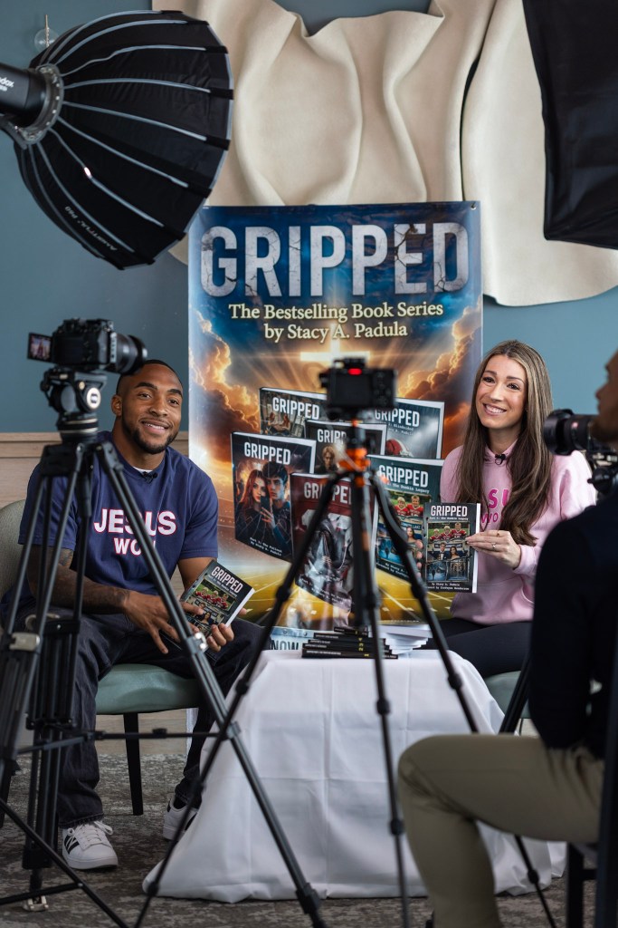 A man and a woman sitting at a table holding books from the 'Gripped' series by Stacy A. Padula, with cameras set up for an interview. A promotional banner for the book series is visible in the background.