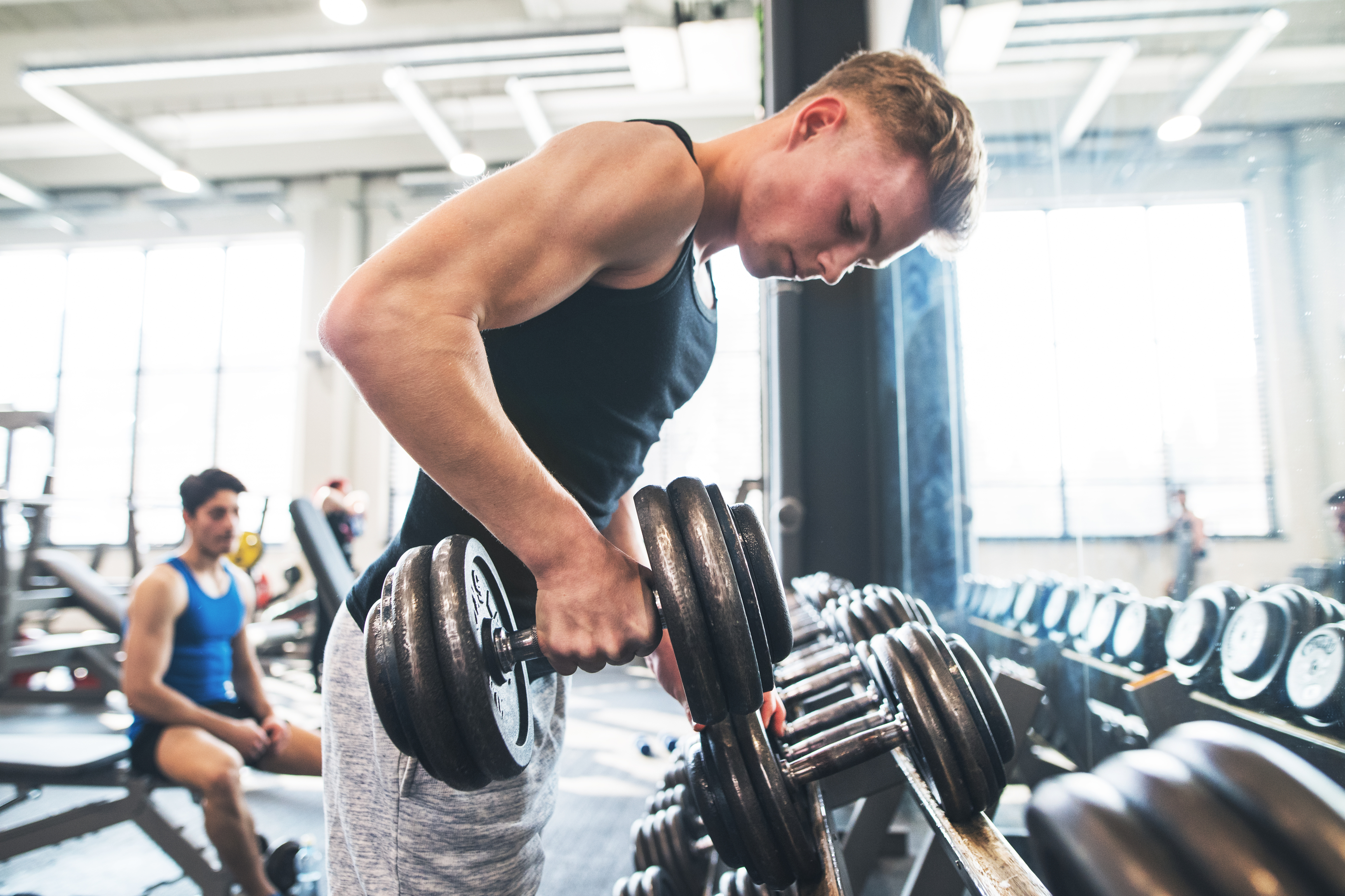 A young man lifting a dumbbell in a gym, focusing on his workout, with another person seated in the background.
