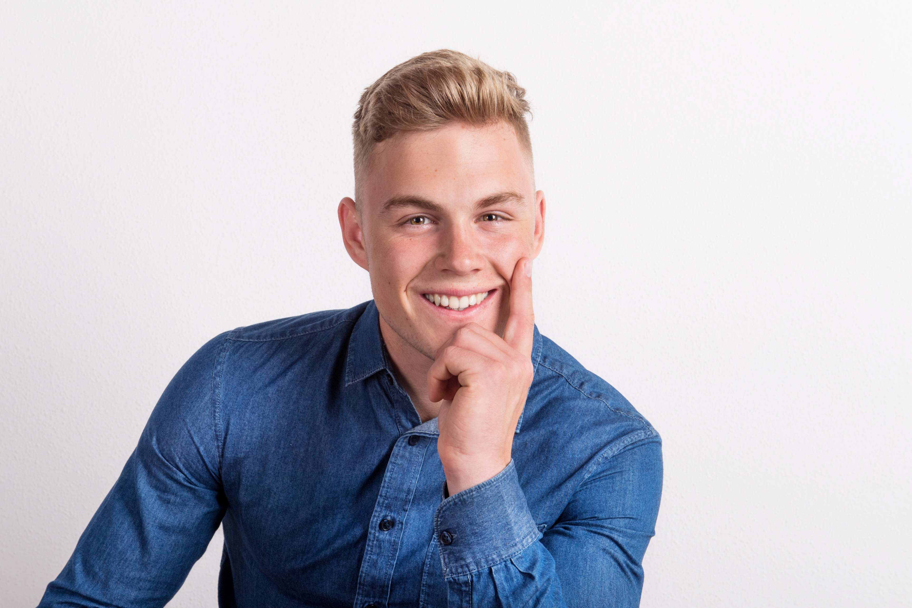 A smiling young man with short blonde hair wearing a denim shirt, resting his chin on his hand against a plain white background.