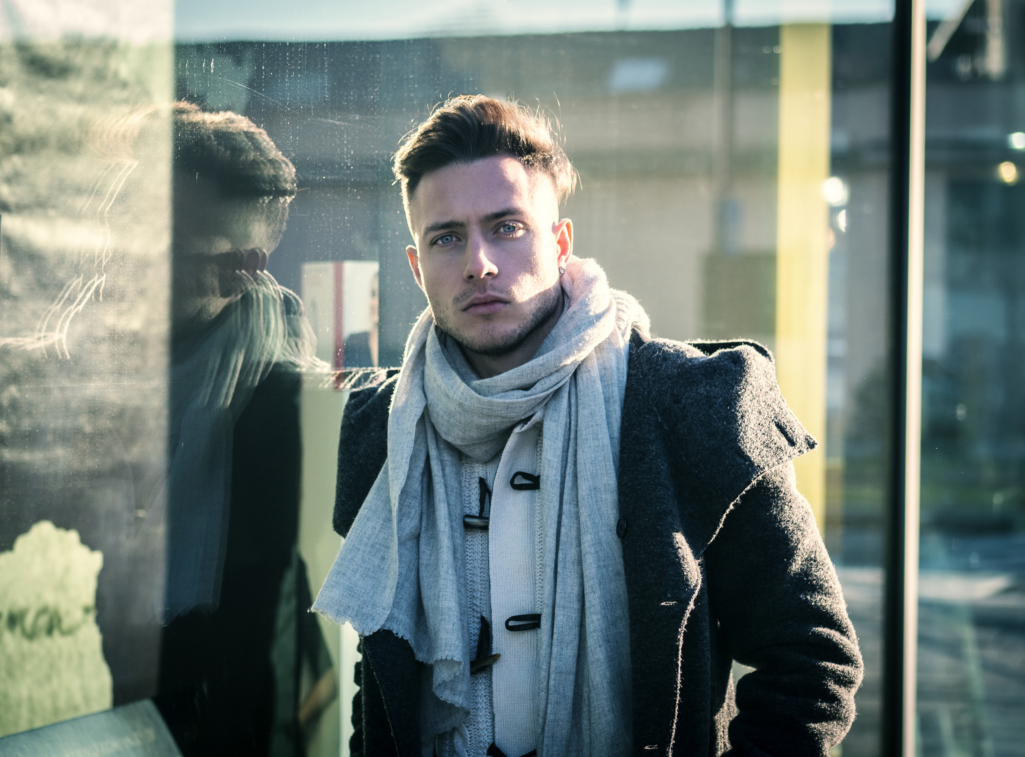 Young man in a gray coat and cream scarf standing next to a reflective surface, looking directly at the camera.
