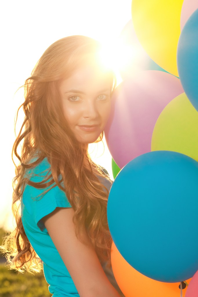 A young woman with long, wavy hair smiles softly while standing among colorful balloons in a sunlit setting.