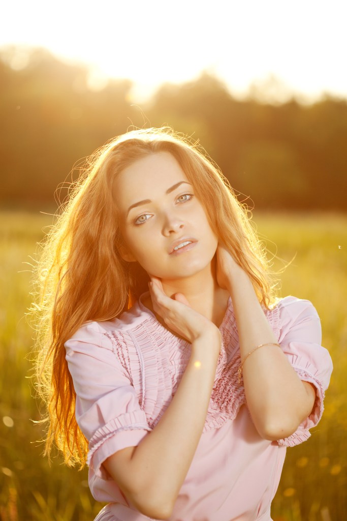 A woman with long, wavy hair posing gently in a sunlit field, wearing a pink blouse and softly touching her neck.