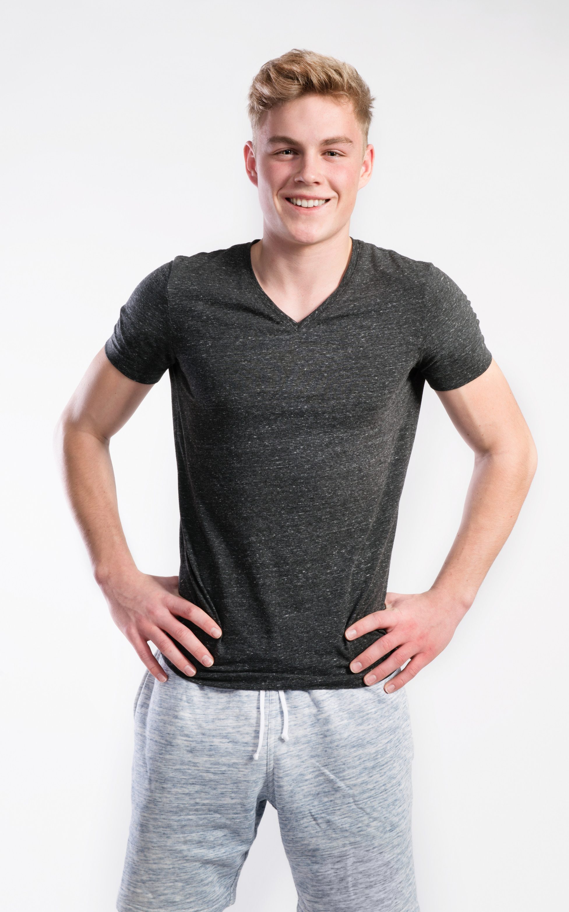 A smiling young man in a gray v-neck t-shirt and light gray shorts, standing with hands on his hips against a white background.