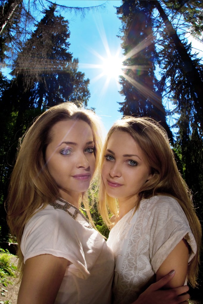 Two young women smiling at the camera in a forest setting with sunlight filtering through the trees.