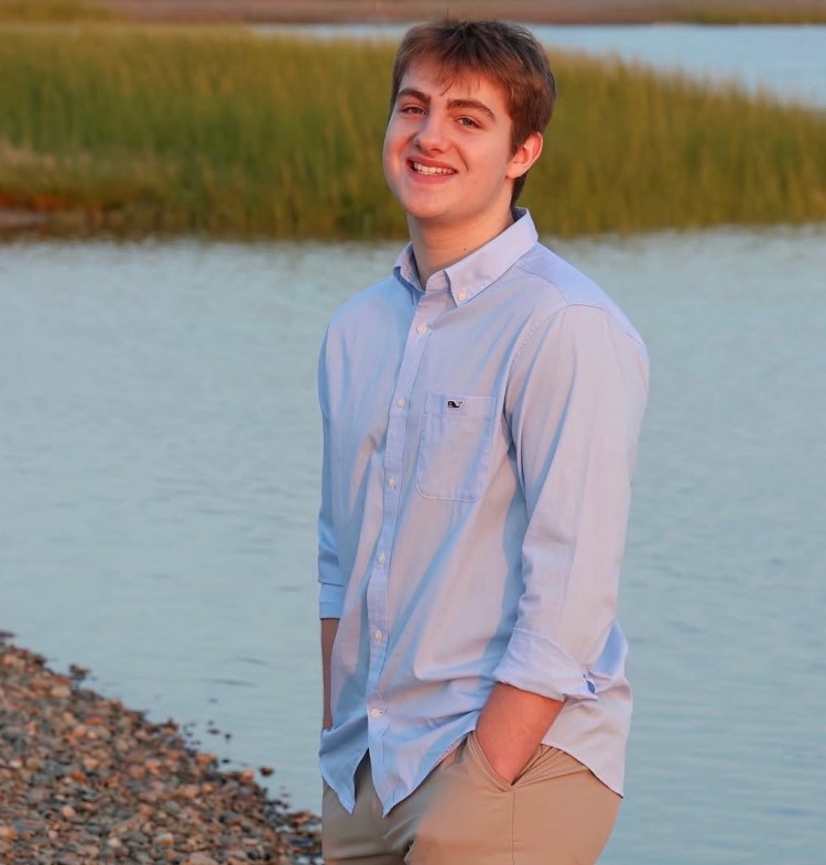 A young man smiling by a water body with green vegetation in the background, wearing a light blue shirt and beige pants.