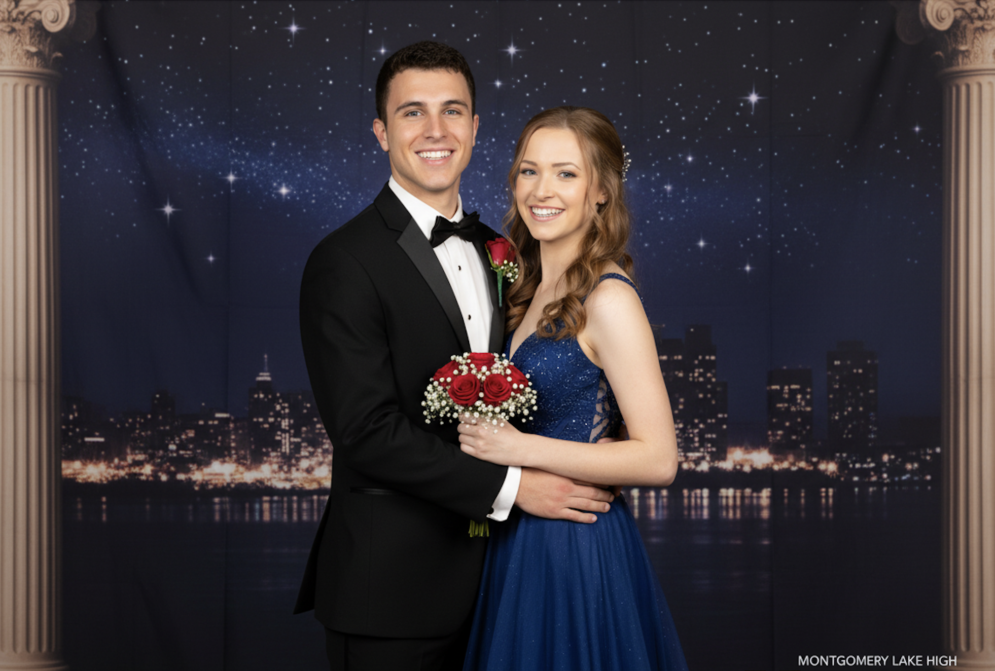 A young man in a black tuxedo poses with a young woman in a blue gown, smiling and holding a bouquet of red roses against a starry night backdrop.